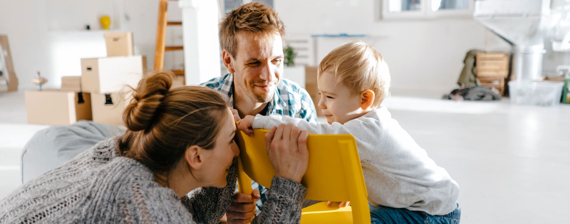 Junge Familie in einem modernen noch nicht fertig eingerichteten Raum. Die Mutter hockt vor einem gelben Stuhl und versteckt ihr Gesicht vor dem Kleinkind, was auf dem Stuhl sitzt.