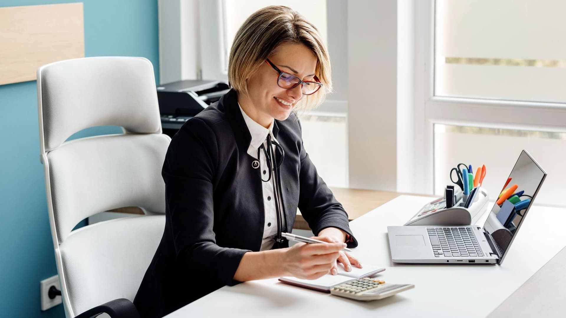 Eine lächelnde Geschäftsfrau mit Brille schreibt in ein Notizbuch an einem hellen Bürotisch mit Laptop und Büromaterialien.