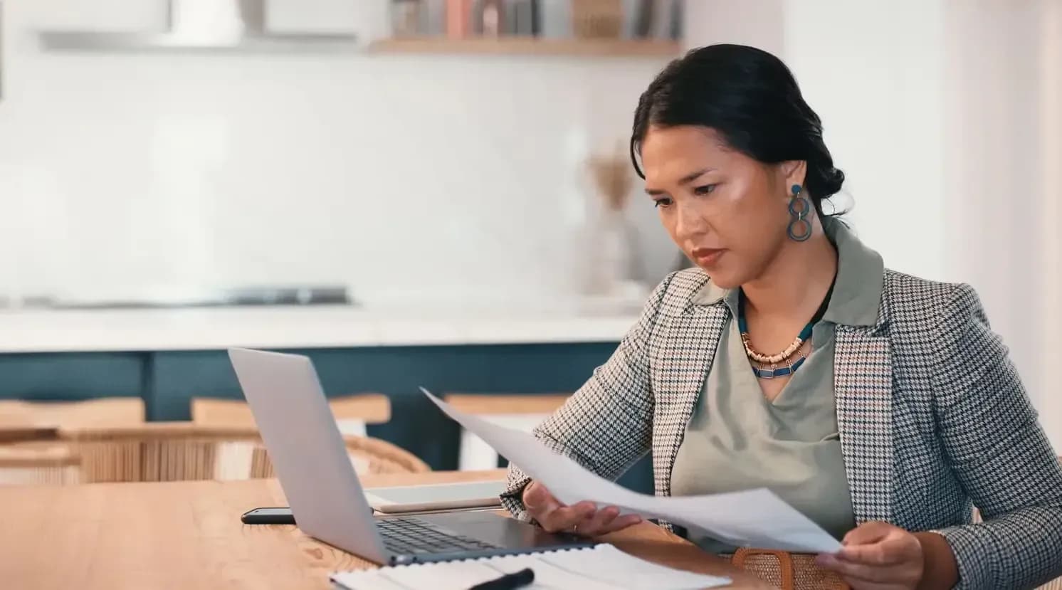 frau sitzt am tisch mit laptop und liest unterlagen in moderner wohnküche