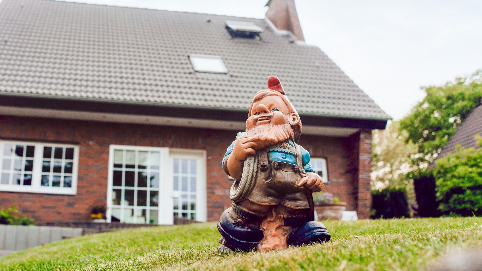 Aufnahme eines Gartenzwergs, der auf einer grünen Wiese vor einem Einfamilienhaus steht.