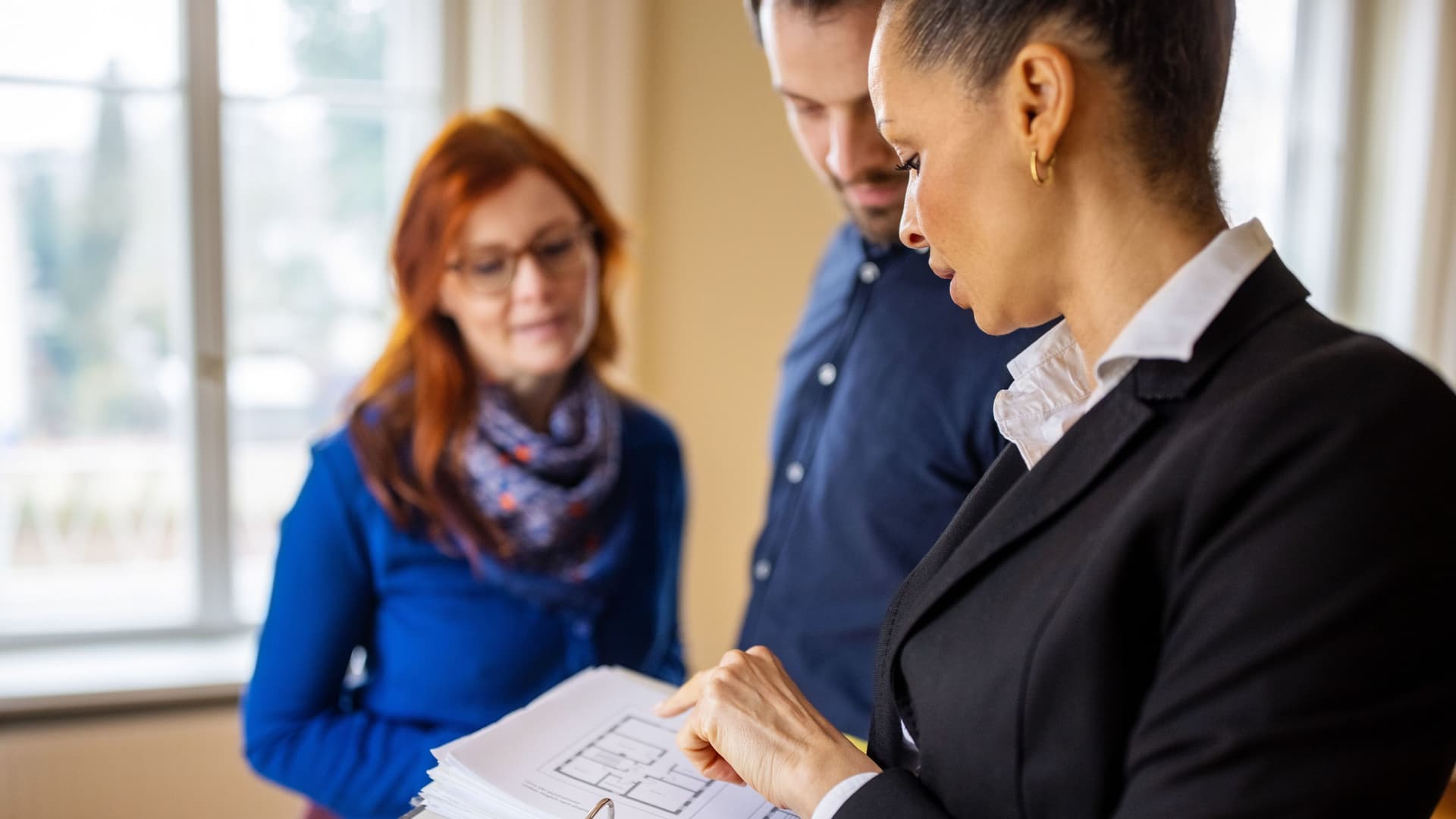 Maklerin zeigt einem Paar eine leere Wohnung anhand von Bauplänen.