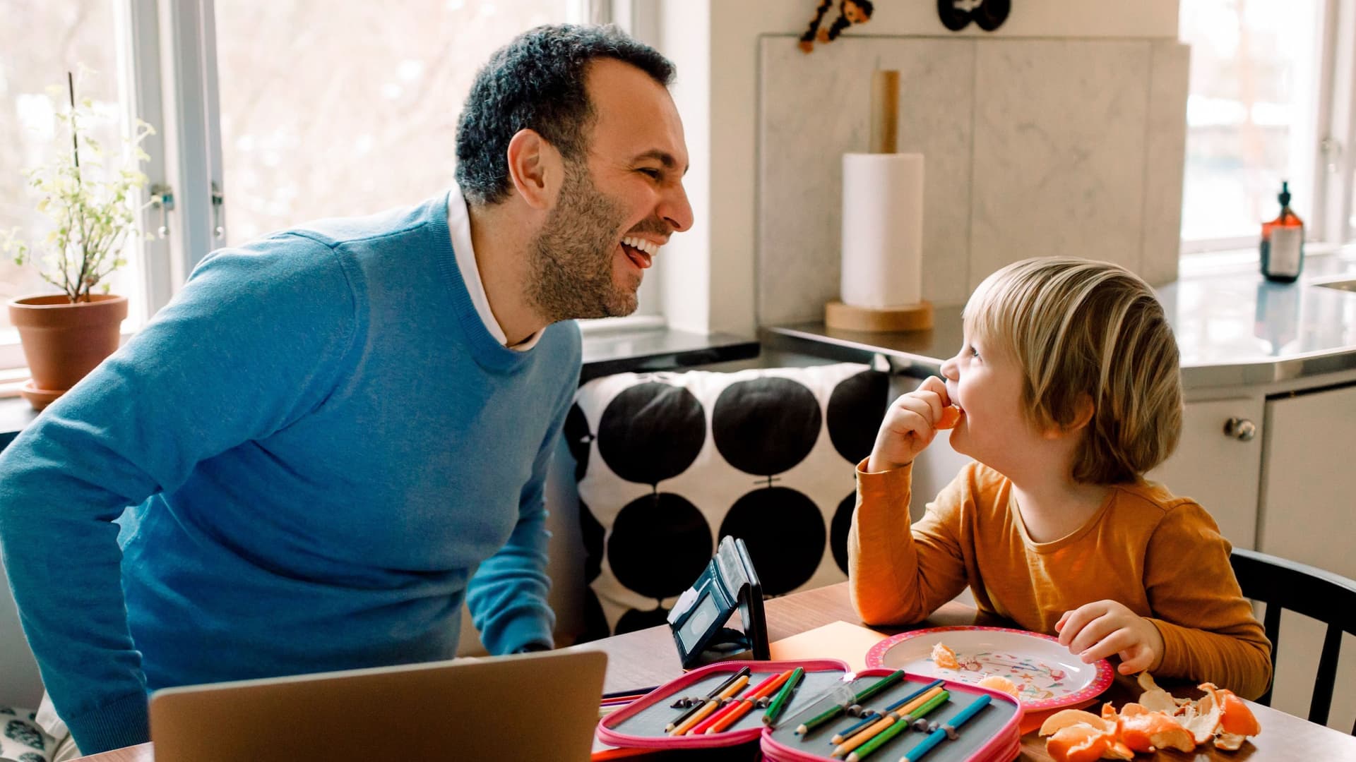 Ein Mann sitzt an seinem Laptop zusammen mit seinem Sohn in der Küche und lacht ihn an.